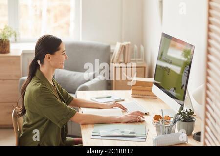 Warm-toned side view portrait of smiling young woman using computer while studying online at home, copy space Stock Photo