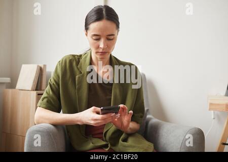 Portrait of modern adult woman using smartphone while relaxing in armchair t home, copy space Stock Photo