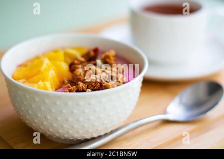 Healthy summer breakfast of a pink smoothie bowl made with frozen banana and raspberry garnished with granola and fresh orange. Stock Photo