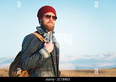 Portrait of a thoughtful stylish hipster with a beard in sunglasses and a hat with a backpack on the background of the snow-capp Stock Photo