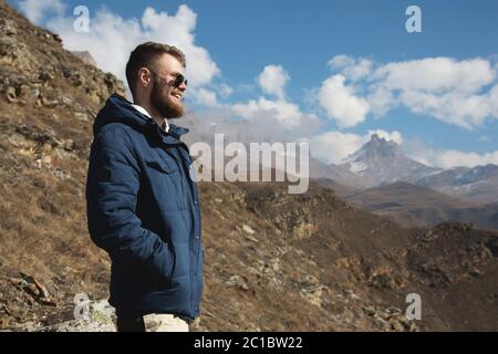 Hipster traveler in a down jacket and sunglasses stands on a mountain slope against the backdrop of epic rocks and smiles. The c Stock Photo