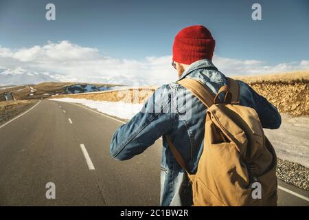 A bearded hipster with an old-fashioned vintage backpack wearing sunglasses with a red hat and jeans jacket and jeans stands on Stock Photo