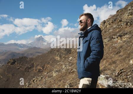 Hipster traveler in a down jacket and sunglasses stands on a mountain slope against the backdrop of epic rocks and smiles. The c Stock Photo