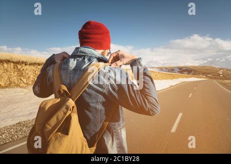 A bearded hipster with an old-fashioned vintage backpack wearing sunglasses with a red hat and jeans jacket and jeans stands on Stock Photo