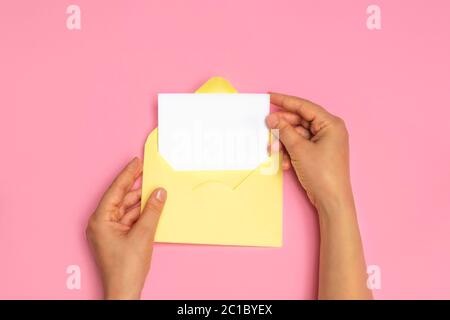 Top view of woman hands holding empty white card and open yellow envelope letter on pink background, mockup Stock Photo
