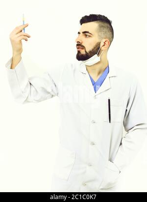 Dentist with beard holds syringe and looks at it attentively. Ambulance service concept. Medicine and recovery idea. Man with assured face in white coat and surgical mask isolated on white background Stock Photo