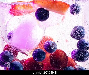 Close up view of the cold and fresh lemonade with bright berries and ice cubes in neon light. Texture of cooling summer's drink with macro bubbles on glass. Fizzing or floating up to top of surface. Stock Photo
