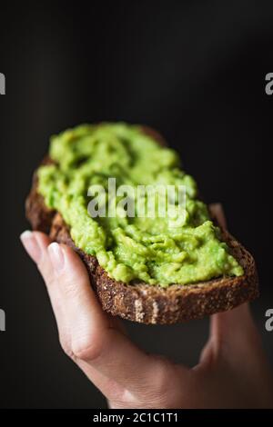 Hand holding rye bread toast with mashed avocado over black background. Healthy eating, vegan vegetarian diet concept. Healthy nutrition Stock Photo