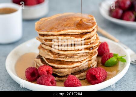 Syrup pouring on stack of whole wheat pancakes served with fresh raspberries on a plate. Closeup view. Healthy breakfast food Stock Photo