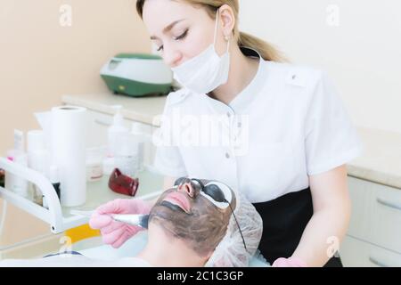 The cosmetologist in pink gloves with a brush applies a carbon mask for peeling on the face of a young girl in a cosmetology roo Stock Photo