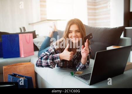 Online shopping, home delivery. Woman lies on a sofa and buys things on the Internet. She is looking at the camera and holding a thumb up and a credit Stock Photo