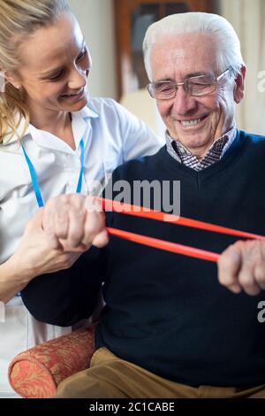 Female Physiotherapist Helping Senior Man To Use Resistance Band At Home Stock Photo