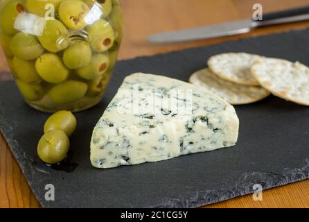 Blue veined cheese on slate with jar of stuffed olives - Stilton or Roquefort type cheese close up o Stock Photo