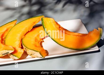 Honey melon or cantaloupe Cucumis melo. Sliced of Japanese melon on white plate with shadows. Summertime fruits.  Stock Photo