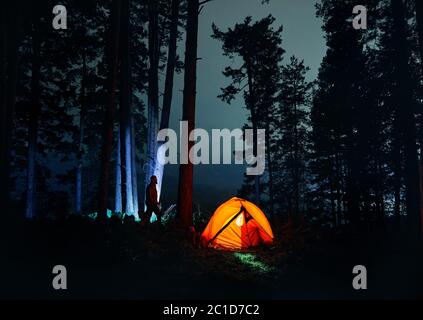 Bearded Man silhouette is walking against tree trunk in the night forest near glowing orange tent in the mountains. Stock Photo