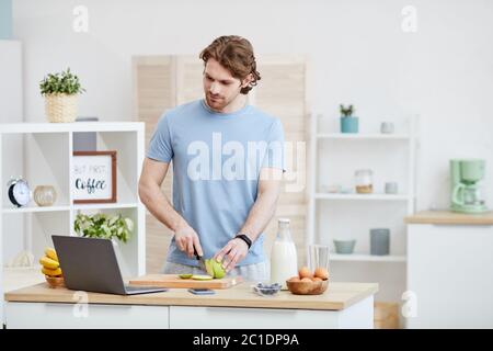 Young man cutting fruit on cutting board and looking at monitor of laptop he cooking using online course at home Stock Photo