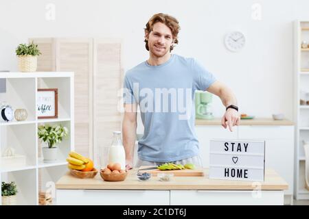 Portrait of young man smiling at camera and pointing at placard with text while cooking in the kitchen Stock Photo