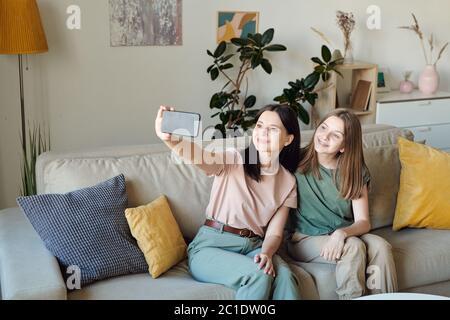 Smiling mother sitting on sofa in living room and taking selfie with daughter on smartphone Stock Photo