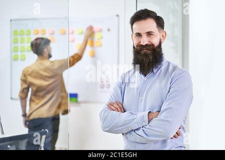Happy smiling bearded senior developer or manager in modern IT office. Another professional working with scrum desk on background. Successful Stock Photo