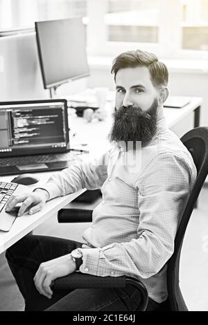 Cheerful senior programmer developer working in IT office, sitting at desk and coding, working on a project in software development company or startup Stock Photo