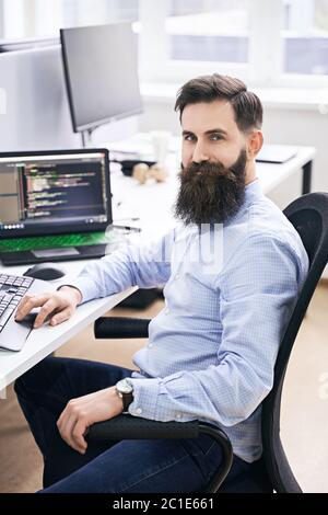 Cheerful senior programmer developer working in IT office, sitting at desk and coding, working on a project in software development company or startup Stock Photo