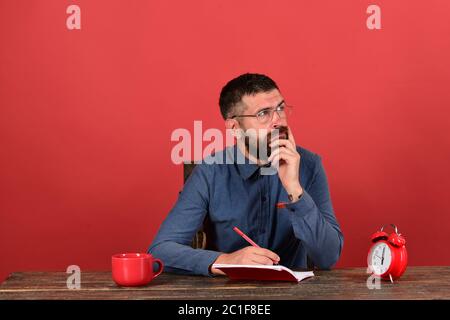 Man with thinking face sits at wooden table. Man with beard and glasses writes in notebook on red background. Exam and studying concept. Cup, retro clock and red book on vintage table Stock Photo