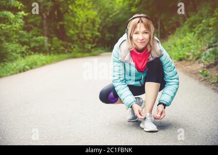 Portrait of a sporty blonde girl in headphones on a run in the forest. A girl sitting tied shoelaces on cross-country shoes. Act Stock Photo