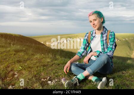 Portrait of young smiling woman traveler with multi-colored hair. Sitting high in the mountains in the evening with a decline in Stock Photo