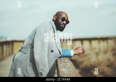 Portrait of a handsome bearded bald African man in sunglasses and coat leaning on a wooden fence outdoors, selective focus, shallow depth of field Stock Photo