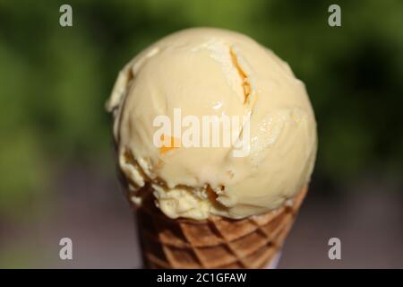 Ball of orange ice cream on top of a waffle cone. Delicious summer season snack usually enjoyed outdoors. Soft green park background. Refreshing! Stock Photo