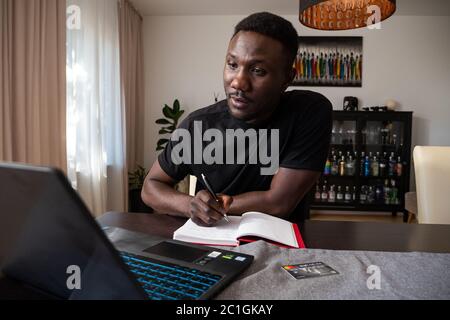 Young black man taking pen and paper notes from laptop seated in living room. Medium shot. Stock Photo