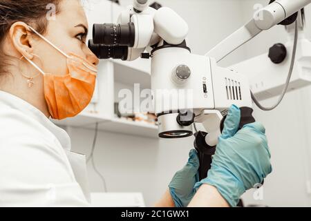Young woman dentist doctor looks through a professional microscope in a dental clinic. A doctor in a disposable medical mask and cap. Advanced Stock Photo