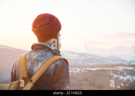 Bearded tourist hipster man in a hat with a backpack stand back on a roadside bump and watching the sunset against the backgroun Stock Photo