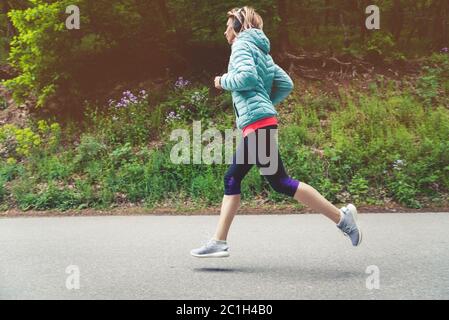 A young blonde woman running is practicing outdoors in a city mountain park in the forest. Warm rays through the branches of tre Stock Photo