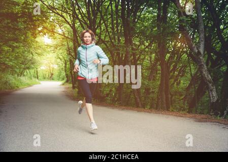 A young blonde woman running is practicing outdoors in a city mountain park in the forest. Warm rays through the branches of tre Stock Photo