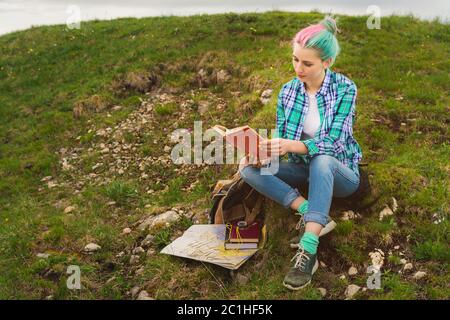 A traveler girl sits in the mountains on the grass and reads a book on the background of epic mountains. The concept of reading Stock Photo