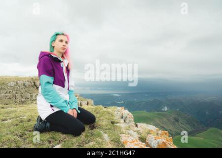 A girl-traveler with multicolored hair sits on the edge of a cliff and looks to the horizon on a background of a rocky plateau Stock Photo