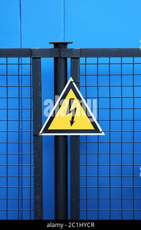 High voltage triangle warning sign mounted on a metal lattice fence on a blue background. Stock Photo