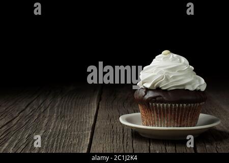 A chocolate cupcake in a typical fluted cupcake liner, topped with whipped cream It is standing on a small ceramic saucer, on weathered wood planks. Stock Photo
