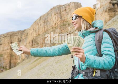 A traveler girl wearing a hat and sunglasses holds a hundred dollar bills in the hands of a fan against the backdrop of rocks on Stock Photo
