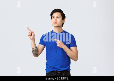 Dreamy young male asian student, guy with aspirations looking and pointing upper left corner with enthusiastic, pleased expression. Taiwanese man in Stock Photo