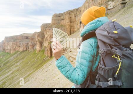 A traveler girl wearing a hat and sunglasses is holding a hundred dollar bills in the hands of a fan against the backdrop of cli Stock Photo