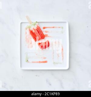 Broken fruit ice cream lolly on a white plate with a pattern from the thawing ice cream on a gray marble background. Copy space Stock Photo