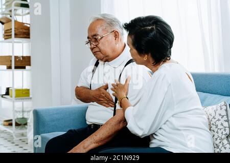 Senior woman taking care of elderly Asian man suffering from heartburn while sitting on sofa in cozy living room at home. Old pensioner sick patient Stock Photo