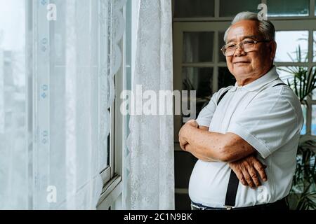 Positive plump senior Asian male smiling for camera and crossing arms while resting near window in cozy room at home Stock Photo