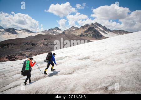 Two tourists, a man and a woman with backpacks and crampons on their feet walk along the glacier against the background of the m Stock Photo