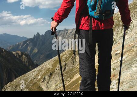The body of man with a backpack and trekking poles stands on top of a rock against the background rocky valley high in the mount Stock Photo