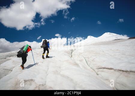 Two tourists, a man and a woman with backpacks and crampons on their feet walk along the glacier against the background of the m Stock Photo