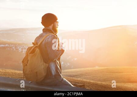 Bearded tourist hipster man in a hat with a backpack is sitting back on a roadside bump and watching the sunset against the back Stock Photo