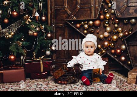 Smiling lovely baby girl in cute dress with headband sitting on bench with lots of christmas presents. Stock Photo